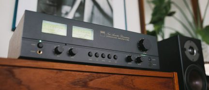Črn stereo hi-fi ojačevalnik NAD C 3050 z retro merilniki na sprednji plošči, postavljen na leseno omarico blizu zvočnika