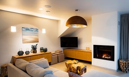Modern, cozy living room with a large copper pendant light above a raw wood coffee table and a lit designer fireplace