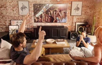 Fekete soundbar házimozi-rendszer mélyládákkal nagy képernyős TV alatt, ipari stílusú, téglafalas nappali, barátok a kanapén