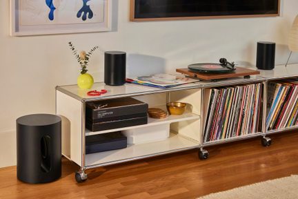Čierny valcový bezdrôtový subwoofer položený na zemi pri bielom hifi nábytku na kolieskach