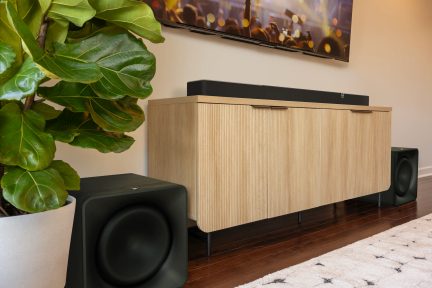 Impianto home cinema con soundbar nera su un mobile TV scanalato color legno chiaro, affiancato da due subwoofer neri