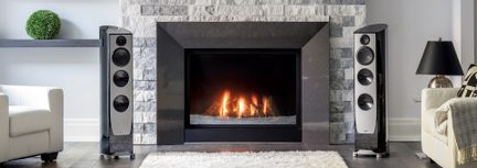 Moderní obývací pokoj pohled zepředu, centrální krb z světle šedého kamene s černým rámem, v němž hoří oheň, po stranách dva sloupové reproduktory v černém laku s hliníkovým předním panelem vybavené čtyřmi kruhovými reproduktory, krémové křesla a černé lampy po obou stranách na tmavé podlaze z parket