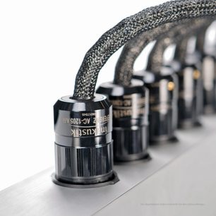 Zbliżenie, widok ukośny, na czarne, błyszczące plastikowe złącza cylindryczne i kable w oplotach w kolorze ciemnoszarym ułożone na prostokątnej, srebrzystoszarej obudowie z szczotkowanego aluminium wielokrotnej listwy zasilającej Inakustik AC-3200 Filter Digital SCHUKO.