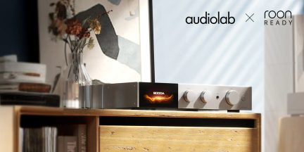 An amplifier with a display showing "9000A" and three circular buttons on a wooden shelf, transparent vase with flowers