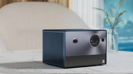 Pelēks taisnstūrveida 4K projektors uz balta galda, skats no trīs ceturtdaļām priekšpuses, redzama lēca un sensori