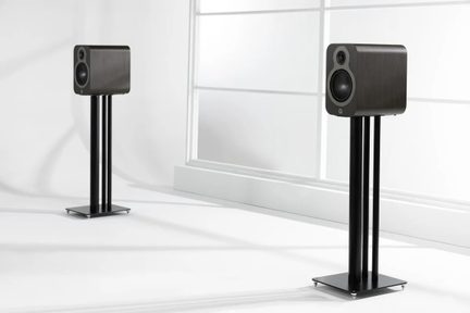 Pair of compact rectangular speakers in dark wood with a black front panel, equipped with circular tweeter and woofer, mounted on black triple tubular metal stands, three-quarter view in a white setting