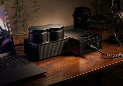 Amplificatore DAC per cuffie nero in metallo, jack e cavo intrecciato, box adiacente con due grandi manopole, scrivania in legno