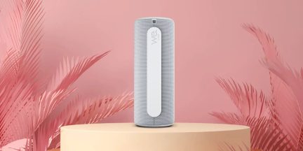 Tragbarer Bluetooth-Lautsprecher We Hear in Weiß im zylindrischen Format auf beigefarbenem Sockel vor rosafarbenem Hintergrund mit tropischem Blattwerk