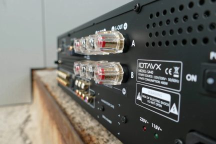 Trebaksvy bakifrån av en svart metallisk ljudförstärkare, närbild på fyra transparenta och guldfärgade högtalarterminaler med röda markeringar, vita tryckta etiketter och en rad cirkulära ventilationsöppningar