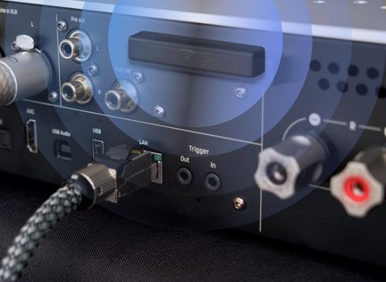 Vedere din spate, în prim-plan, a unei carcase audio din metal negru mat, cu dongle negru dreptunghiular având un pictogramă wifi, ieșiri RCA pre-out și sub-out, port USB, port LAN ocupat de un cablu împletit, mini-jack-uri trigger și borne pentru boxe roșii și gri