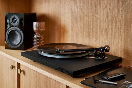 Platine noire sur étagère en bois clair, disque vinyle noir sur plateau en verre, bras de lecture, vue de trois-quarts, enceinte et vase