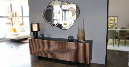 Nábytek s vlnitým dřevěným čelem, černá lampa, dvě černé koule, tři protáhlé vázy, kulaté nástěnné zrcadlo