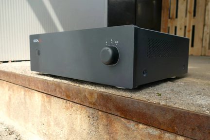 Rektangulær lydforsterker i matt svart metall, sett fra trekvart forfra, med stor rund gradert dreieknapp til høyre, firkantet av/på-knapp til venstre og perforert ventilasjonsgitter på siden