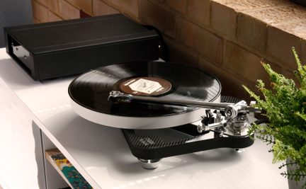 Giradischi con braccio metallico e disco nero su piatto bianco, corpo rettangolare nero sul retro, pianta verde a destra
