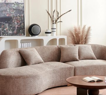 Divano beige curvo con 3 cuscini, tavolino rotondo in legno, scaffale bianco con oggetti decorativi e quadro floreale incorniciato.