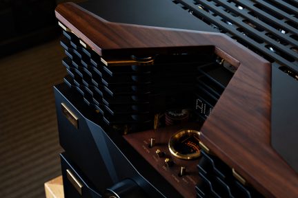 Detailní záběr shora na audio zesilovač s elektronkami v černém a ořechovém provedení, černý kovový kryt s žebrovaným chlazením, horní rámeček z hnědého dřeva, ovládací prvky a obruče VU metru z mosazi, částečně viditelné elektronky a digitální displej