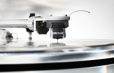 Nærbillede fra siden af en sort og grå phono-pickup i plastik og metal monteret på en sølvfarvet arm, nålen rører den blanke overflade på en vinylplade
