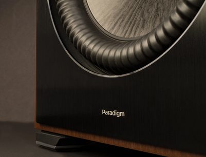 Zblízka čelní pohled na subwoofer Paradigm, světlý dřevěný kužel s koncentrickými drážkami, bílé logo na černé přední straně