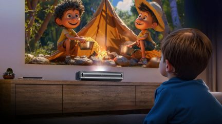 Child sitting, back visible, watching animated projection on wall, projector on wooden furniture, plant and book on the left