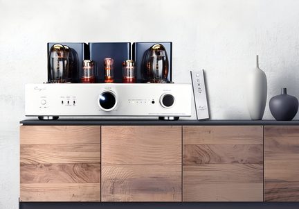 Pohled zepředu na hi-fi lampový zesilovač z leštěného stříbrného kovu, se skleněnými jantarovými lampami seřazenými na vrchní části, dvěma velkými černými knoflíky na přední straně, umístěný na nízké skříňce ze světlého dřeva vedle nakloněného stříbrného dálkového ovladače a dvou matných šedých váz.