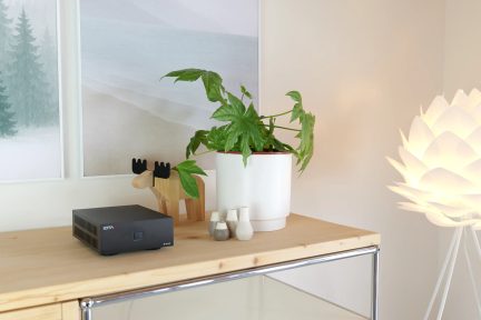 Amplificador IOTAVX preto compacto com botão quadrado na parte frontal e aberturas de ventilação laterais, colocado sobre um móvel claro