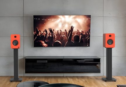 Sort fladskærms-tv monteret på en grå betonvæg, flankeret af to orange, matte kubeformede højttalere med sort, cirkulær enhed, placeret på sorte metalstativer, set forfra