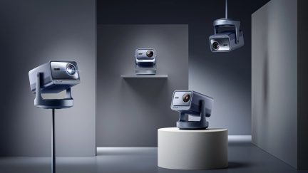 Patru proiectoare gri, poziții variate, pe suporturi și suspendate, într-o cameră cu pereți gri, iluminare blândă