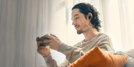 Person sidder, langt hår, sorte hovedtelefoner, beige trøje, holder en smartphone, orange pude, hvide gardiner i baggrunden