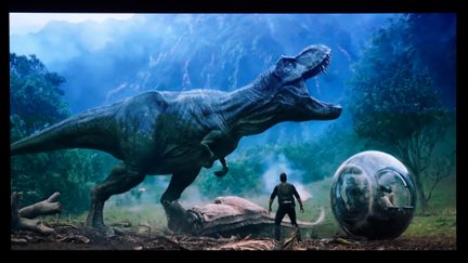 Filmscen i ett brett plan i en dimmig djungel, grågrön dinosaurie sedd från vänsterprofil med öppen mun, man från ryggen i mörk klädsel i mitten och sfärisk genomskinlig kapsel i glas eller plast till höger