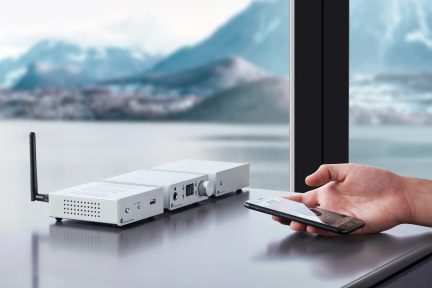 Tři bílé obdélníkové elektronické přístroje zarovnané na hladkém povrchu, vertikální anténa, ruka držící smartphone