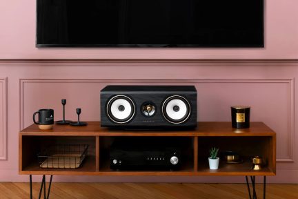 Black Triangle center speaker with dual white drivers, placed on a wooden TV stand beneath a wall-mounted screen