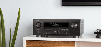 Denon AVC-X6500H sort hjemmebiografsforstærker, set forfra, placeret på et lyst trækabinet med en grøn plante til venstre