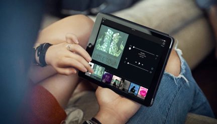Černý tablet držený v levé ruce, na displeji je zobrazena hudební aplikace, pravá ruka se dotýká displeje, viditelné jsou roztrhané džíny