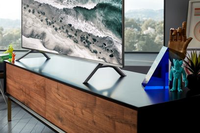 Samsung QE65Q6F 2018 fladskærms-TV, der viser en brydende bølge, stillet på et træmøbel, farverige figurer ved siden af