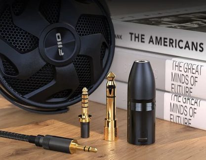 Primo piano, vista obliqua, su un cavo audio mini-jack intrecciato nero con connettore metallico dorato, due adattatori jack metallici dorati e un piccolo contenitore cilindrico in plastica nera opaca, il tutto posato su un vassoio di legno davanti a un altoparlante circolare nero e diversi libri impilati