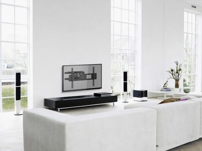 Modernes weißes Wohnzimmer mit Sofa, schwarzem TV-Möbel, Wandhalterung für den Fernseher, Lautsprechern, Pflanzen und großen Fenstern