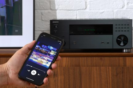 Hlavní smartphone zobrazující obrazovku s přehrávanou hudbou, černý audio přijímač s tlačítkem hlasitosti, zeď z bílých cihel