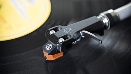 Nahaufnahme, schräge Ansicht, Platin-Tonabnehmerzelle aus mattschwarzem Metall mit zwei silbernen Schrauben und gebogenem Hebel, schwarze und orangefarbene Kunststoffspitze, auf einer schwarzen Schallplatte platziert