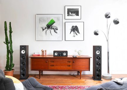 Sala minimalista, vista frontal, com duas colunas de som pretas em madeira ladeando um móvel baixo vintage de madeira que suporta uma coluna central preta, alguns livros, figuras decorativas e um vaso
