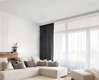 Šviesus salonas, kampinis bežinis audeklinis sofa ir otomanas, taupės pagalvės, baltos tūlės ir pilkas užuolaidas, priekinis vaizdas