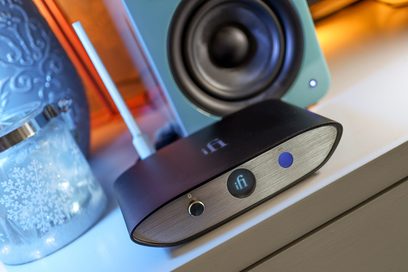 Bezdrátový přenosový systém iFi Audio Zen Blue V2 v černé barvě, pohled zepředu a z profilu, umístěný na bílém stojanu vedle modrého skla