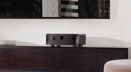 Zwarte metalen audioversterker, vooraanzicht, twee grote knoppen en centraal rond display, op een donkerhouten meubel