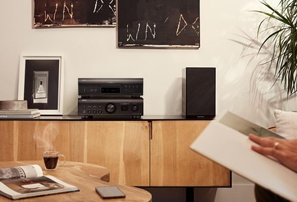 Pohled zepředu na černou kovovou hi-fi soupravu, složenou z CD přehrávače a zesilovače s hladkým předním panelem, displejem a tlačítky, umístěnou na nízkém nábytku ze světlého dřeva vedle černé obdélníkové reproduktorové skříně s látkovou mřížkou.