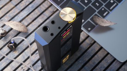 Vista en ángulo inclinado de una caja de audio portátil rectangular negra de aluminio, con laterales acanalados, gran rueda de volumen circular dorada y cuatro tomas de audio alineadas en el borde superior, colocada sobre listones de madera junto a unos auriculares con cable y un teclado de ordenador portátil.