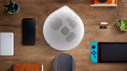 Set fra oven: en hvid, cirkulær højttaler, en sort smartphone, en blå konsol, en USB-nøgle, et kamera, en notesbog, en plante og et batteri.
