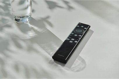Černá prodloužená dálková ovladač Samsung, přední strana, položená na bílém odrazivém povrchu vedle sklenice vody