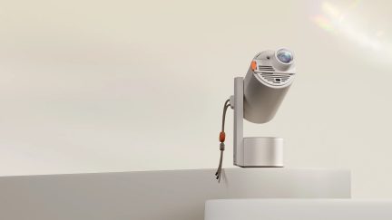 Silver cylindrical projector mounted on a stand, lens facing forward, cord with orange tip, beige background