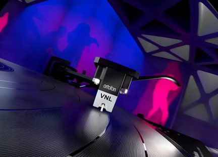 Close-up de uma célula fonocaptora Ortofon VNL preta e cinza, em plástico e metal, sobre um disco de vinil sulcado, vista oblíqua