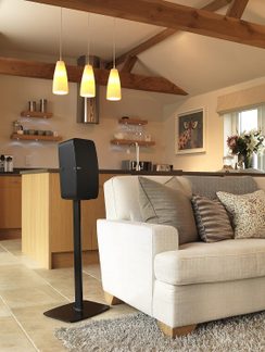 Salotto con divano beige, cuscini decorativi, tappeto grigio, supporto nero con altoparlante, cucina a vista con isola, lampade a sospensione, travi in legno, mensole a muro, lavello in acciaio inox, quadro a parete, vaso con fiori, piastrelle beige