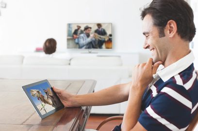 Smilende mand ser på en tablet, der viser et billede af kameler i en ørken, med et fjernsyn i baggrunden